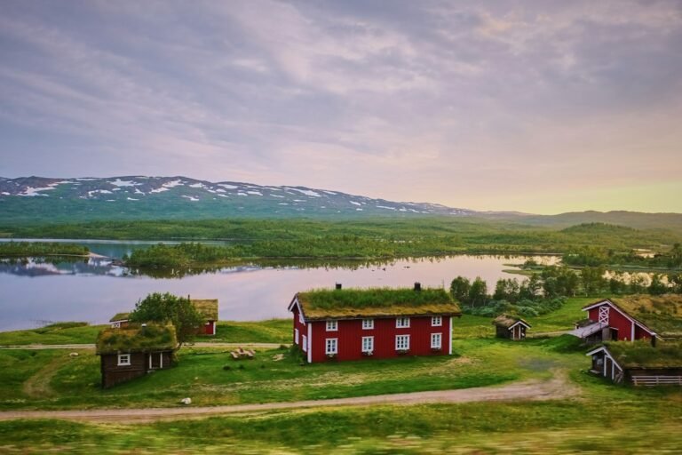 Red wooden cabins with grass roofs by a lake and snowy mountains
